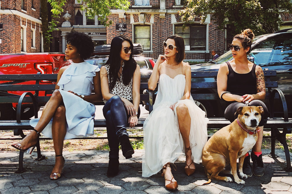 Four women sit on a bench. One of them has a dog sitting by them on the ground.