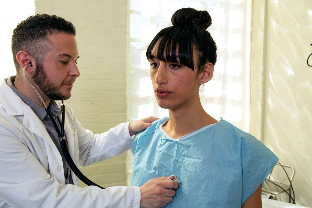 A transgender woman in a hospital gown being treated by a doctor, a transgender man.