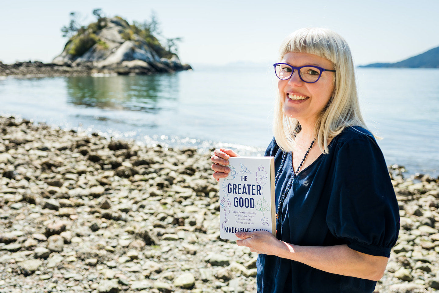 Madeleine Shaw stands on a beach holding a copy of her book, The Greater Good