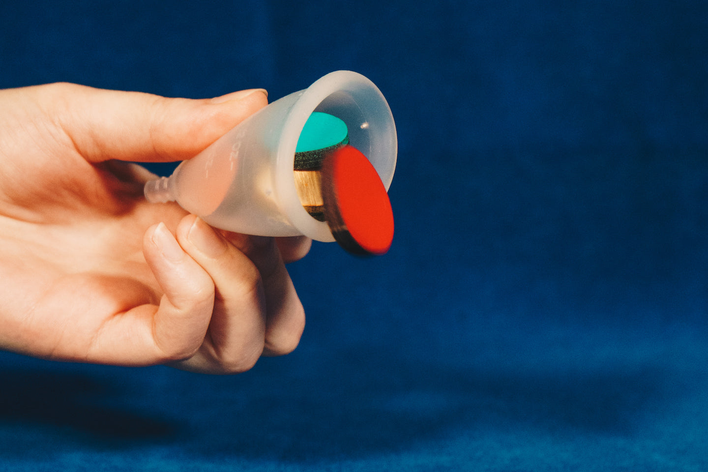 A hand holding a menstrual cup on its side. The cup is filled with coloured shapes, which have started to fall out of it. The background is blue.