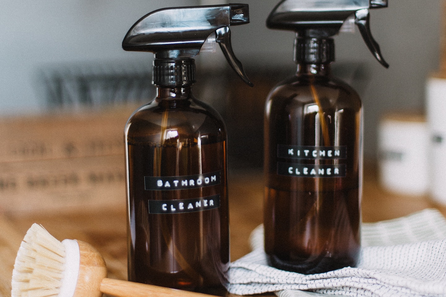 Two homemade cleaners in amber-glass spray bottles sit on a wooden counter. One of the bottles is labeled "Bathroom Cleaner", the other is labeled "Kitchen Cleaner". There is a wooden dish brush laying nearby.