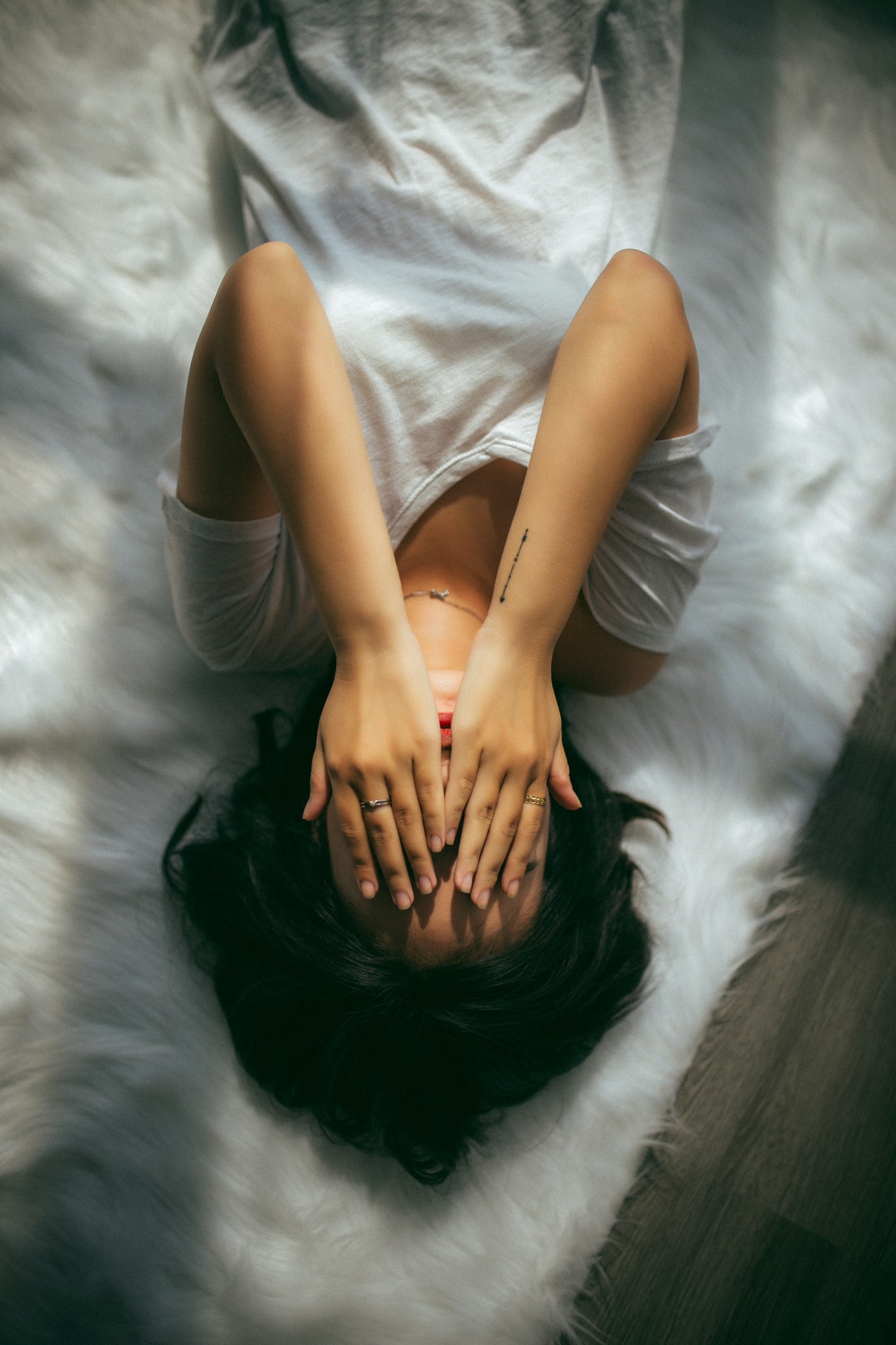 Woman lying on her stomach on a white bed. She's wearing a grey shirt, one arm above her head.